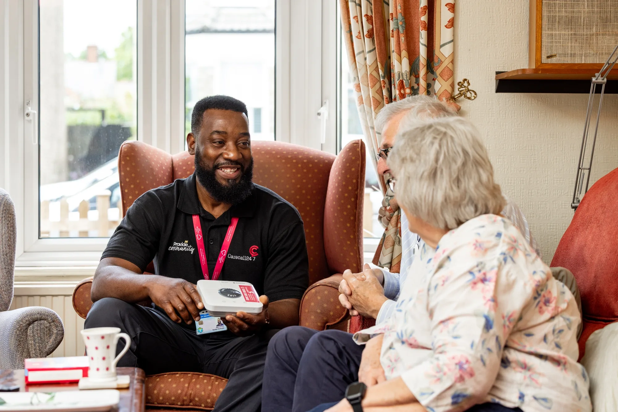 A Provide Carecall247 team member showing a personal alarm to an older couple in their living room.